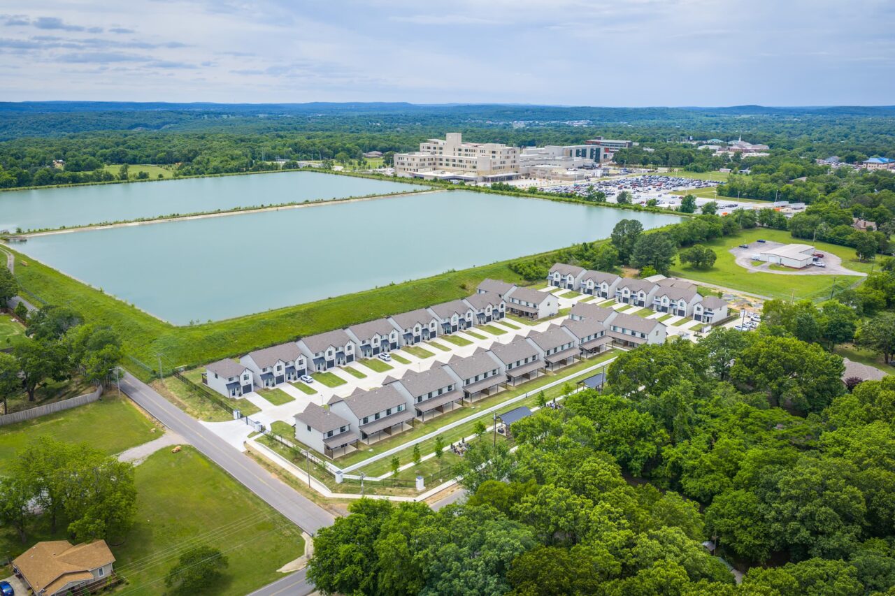 Aerial view of a suburban housing development beside a large rectangular pond and surrounding trees, with a modern campus building and parking lot in the distance.